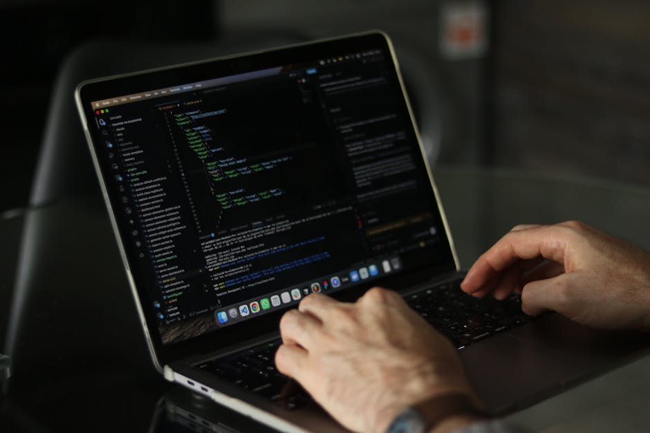 A close-up view of a person's hands typing on a laptop keyboard, with the screen displaying lines of code in a dark-themed editor. The setting appears to be indoors with a polished, reflective surface