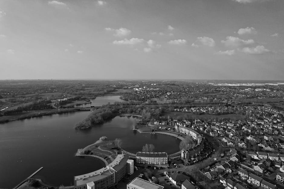 Aerial black-and-white photograph of Feltham TW13, showcasing a serene lakeside residential area with a curved water body surrounded by a mixture of houses, apartment buildings, and some greenery. The