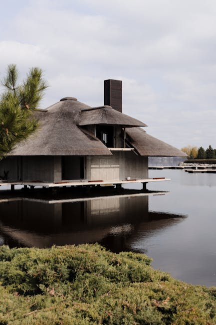 A modern house with a thatched roof situated by a calm body of water, reflected clearly on the surface. Surrounding the house, there are lush green bushes and low-lying plants along the shoreline. The